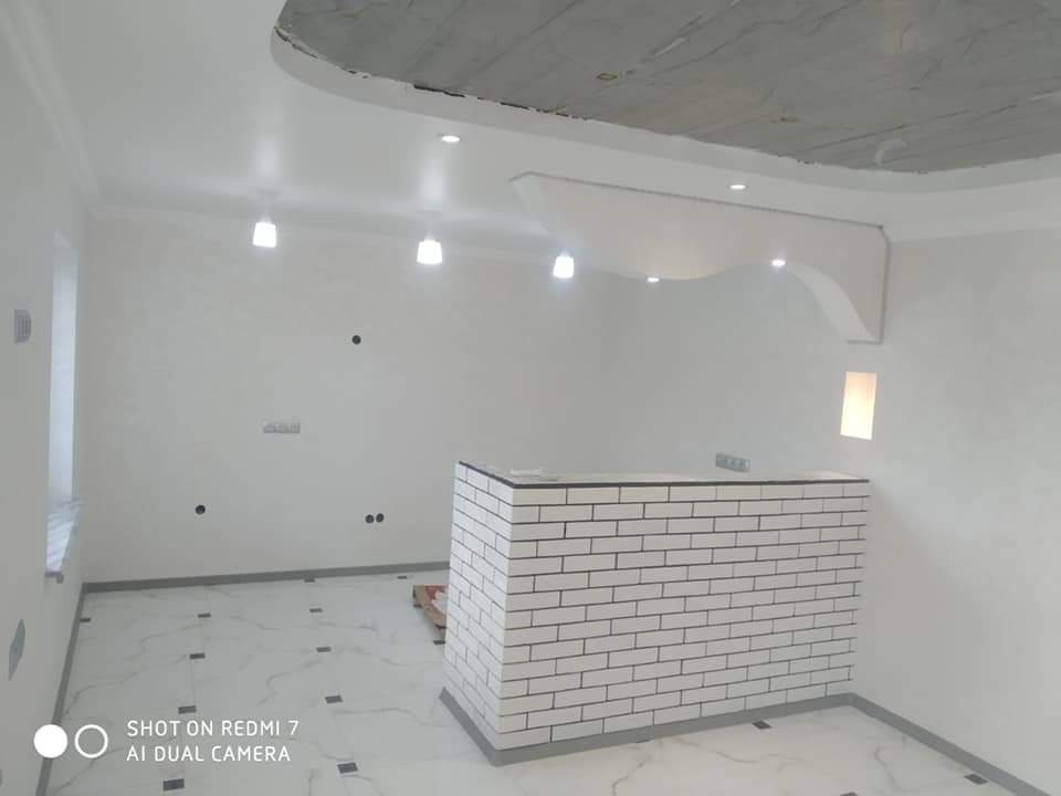 Wnętrze pomieszczenia w trakcie remontu: białe ściany, marmurowa podłoga z czarnymi akcentami, niedokończony sufit z elementami dekoracyjnymi i oświetleniem punktowym, oraz murowana z białej cegły...