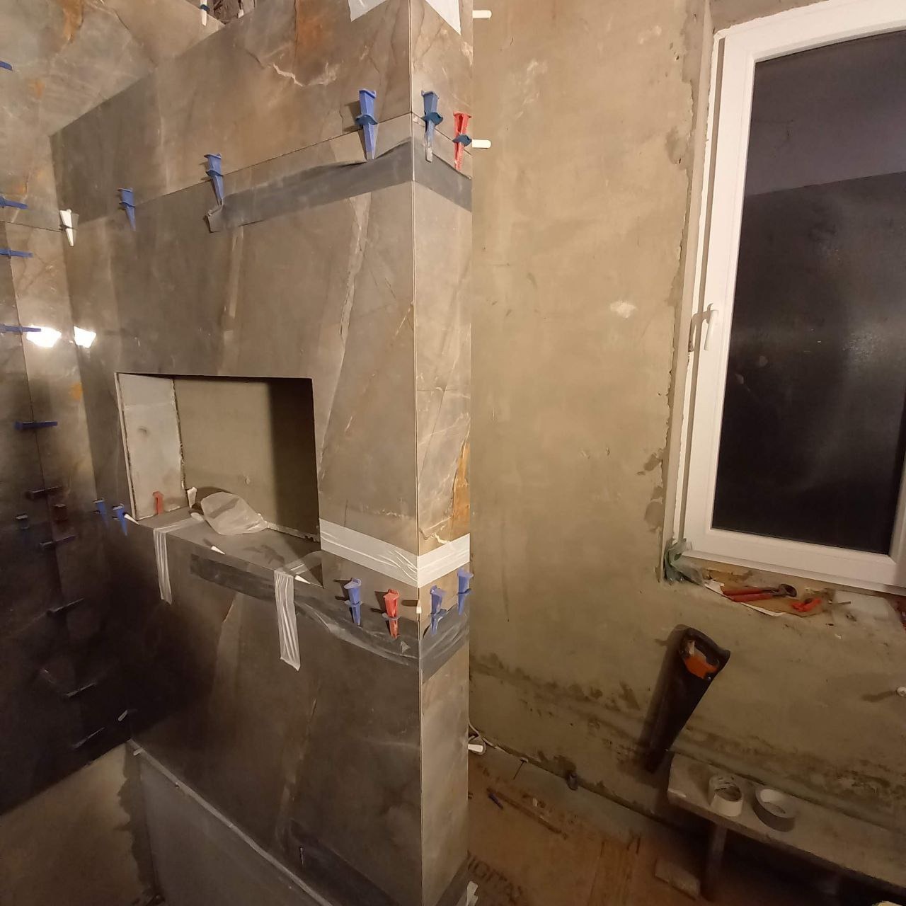 Montaż płytek imitujących marmur w łazience, widoczne krzyżyki dystansowe i taśma maskująca, okno z narzędziami w tle.