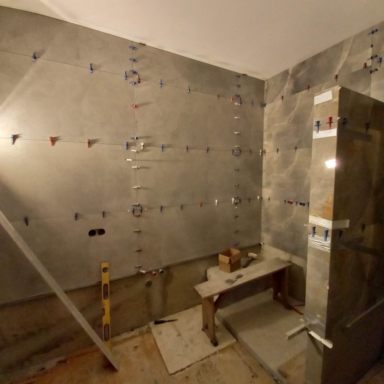 Montaż płytek w łazience z użyciem poziomicy i systemu poziomowania płytek, widoczny fragment kabiny prysznicowej w trakcie obkładania płytkami.