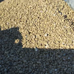 Kopalniany ładunek: pełna przyczepa drobnego, brązowego kruszywa skalnego w słońcu, z widocznymi detalami tekstury i cieniem rzucanym na część materiału, w tle drzewa bez liści i sygnalizacyjne...