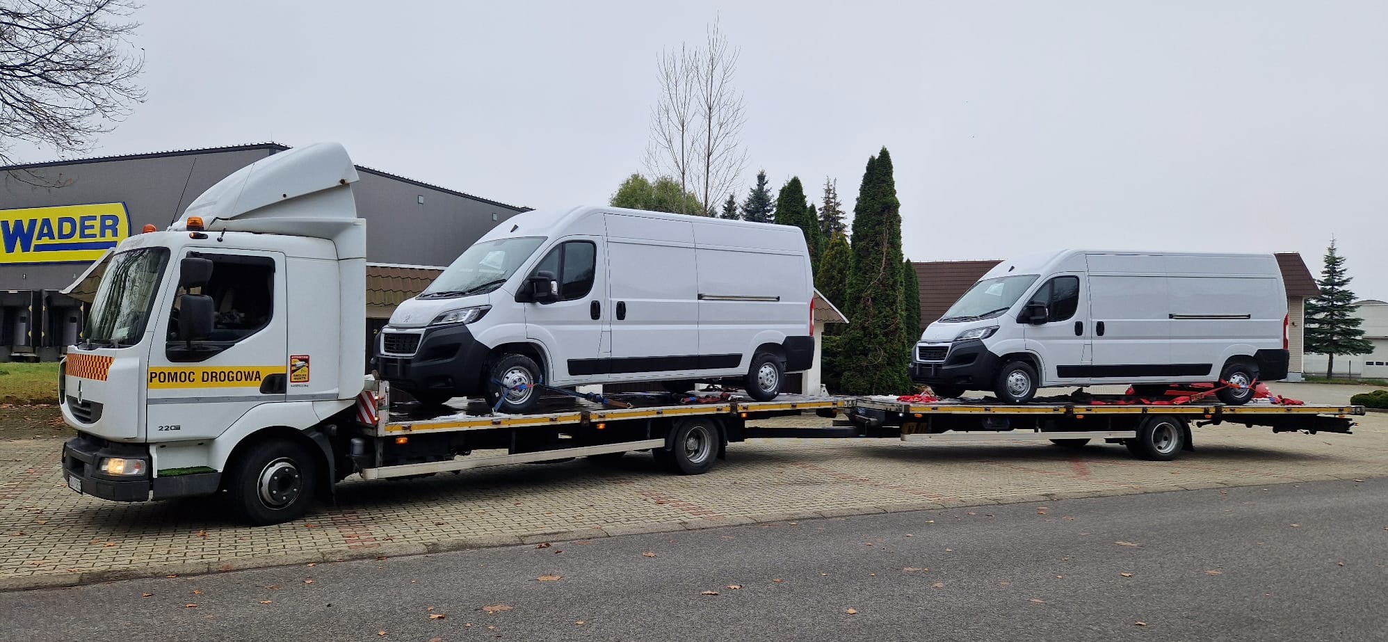 Laweta z dwoma białymi busami zabezpieczonymi pasami transportowymi, widoczne oznaczenie 'Pomoc Drogowa' na kabinie ciężarówki, budynek Wader w tle.