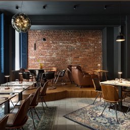 MrMario.pl - Stylowe wnętrze restauracji z odsłoniętą cegłą, ciemnymi akcentami i designerskimi lampami. Ustawione stoliki i krzesła, wzorzysty dywan, okna z żaluzjami.