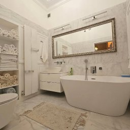 MrMario.pl - Elegancka łazienka w bieli z marmurowymi ścianami. Wolnostojąca wanna, lustro w ozdobnej ramie i szafka z umywalką. Na półkach ułożone ręczniki.