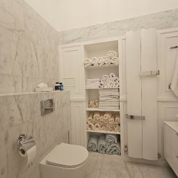 MrMario.pl - Elegancka łazienka w bieli z marmurowymi ścianami i podłogą. Podwieszana toaleta, szafka z półkami na ręczniki i minimalistyczny design.