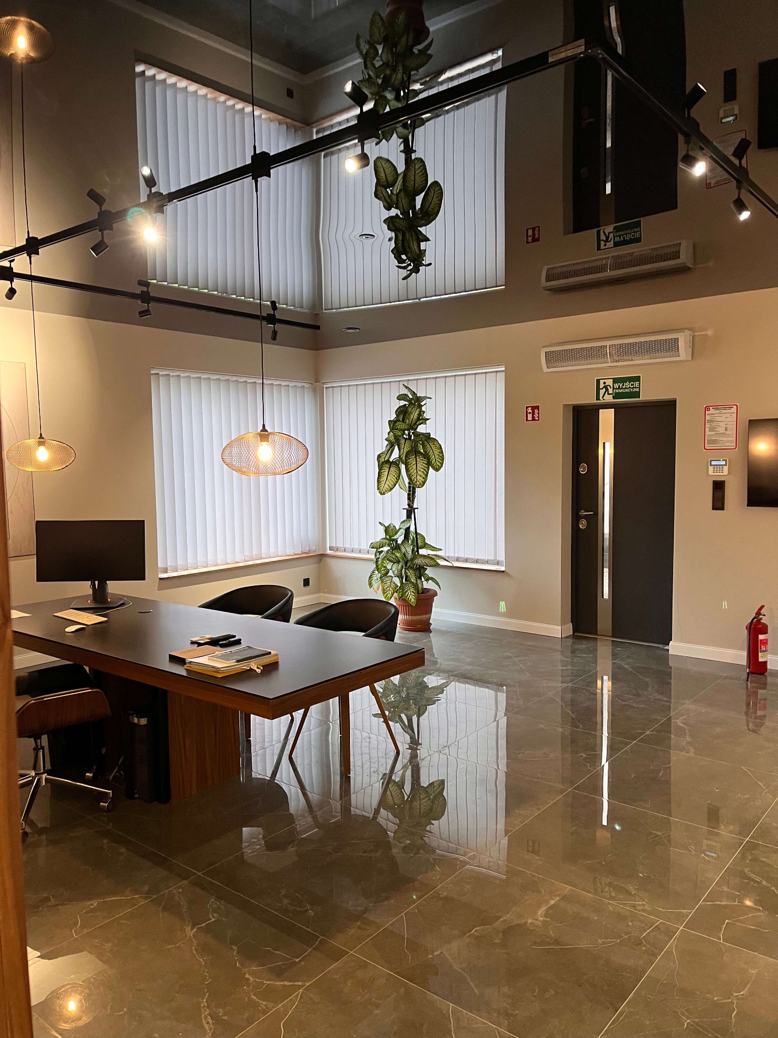 Eleganckie wnętrze biurowe z błyszczącą, marmurkową podłogą, nowoczesnym oświetleniem i minimalistycznym wystrojem. Widoczny stół konferencyjny i rośliny doniczkowe.