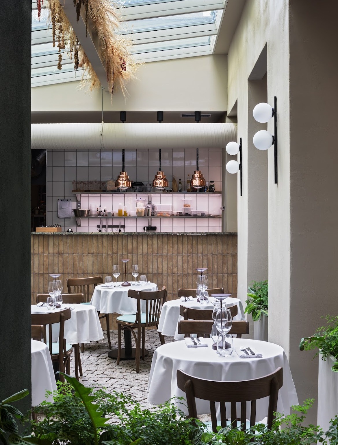 Stylowe wnętrze restauracji z otwartą kuchnią, okrągłymi stolikami nakrytymi białymi obrusami i dekoracyjnym oświetleniem. Widoczne elementy wykończenia ścian i podłogi.