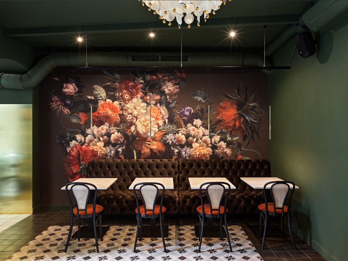 Wnętrze lokalu: ciemnozielone ściany, tapeta w kwiaty, pikowana sofa pod ścianą, białe stoliki, krzesła z wiklinowym oparciem, wzorzysta podłoga i ozdobny żyrandol.