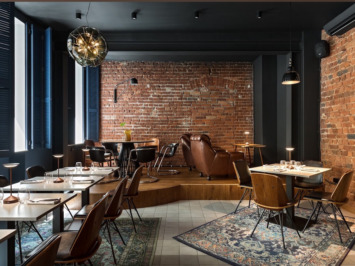 Stylowe wnętrze restauracji z odsłoniętą cegłą, ciemnymi akcentami i designerskimi lampami. Ustawione stoliki i krzesła, wzorzysty dywan, okna z żaluzjami.