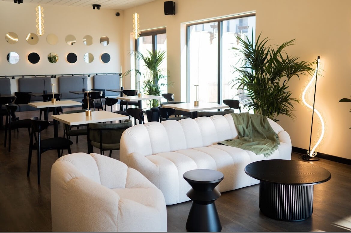 Eleganckie wnętrze kawiarni: biała sofa z zielonym pledem, stoliki z czarnymi krzesłami, okrągłe lustra na ścianie i wysokie rośliny doniczkowe.