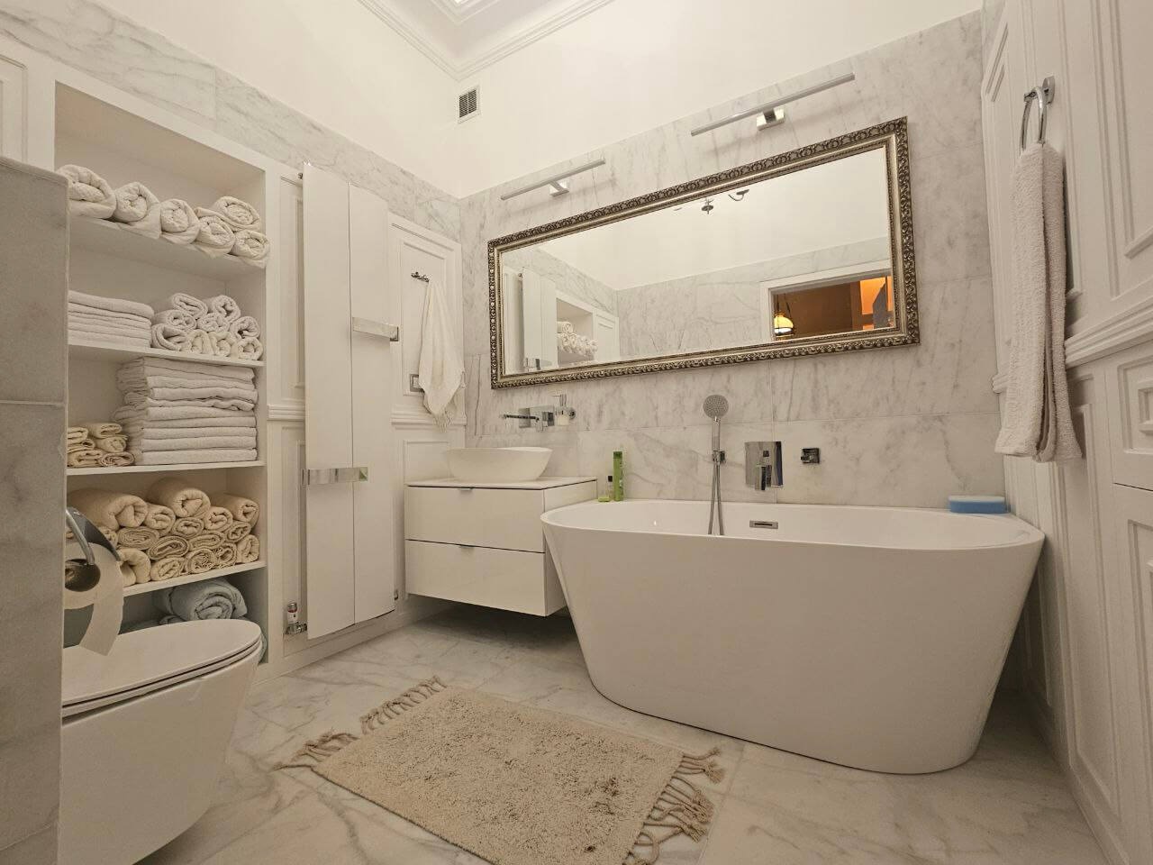 Elegancka łazienka w bieli z marmurowymi ścianami. Wolnostojąca wanna, lustro w ozdobnej ramie i szafka z umywalką. Na półkach ułożone ręczniki.