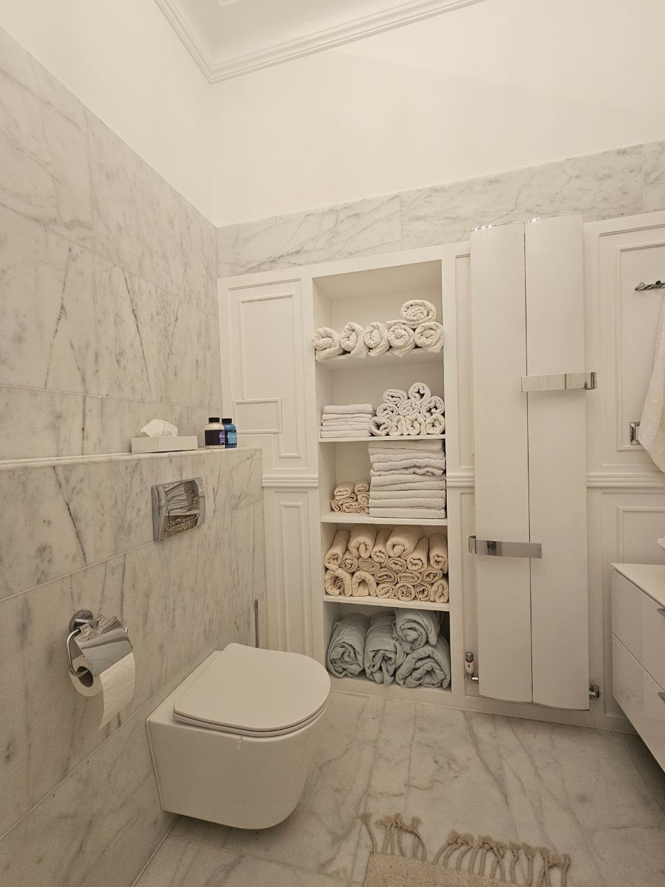 Elegancka łazienka w bieli z marmurowymi ścianami i podłogą. Podwieszana toaleta, szafka z półkami na ręczniki i minimalistyczny design.