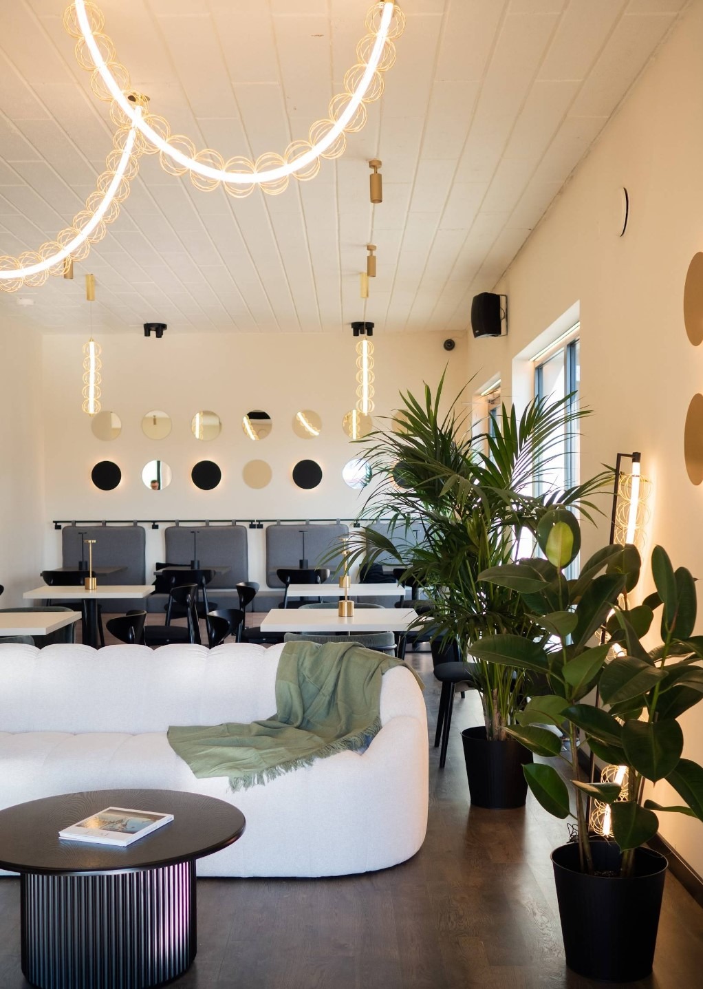 Stylowe wnętrze kawiarni z białym narożnikiem, roślinami doniczkowymi i oryginalnym oświetleniem. Okrągłe lustra i lampy wiszące dodają elegancji.