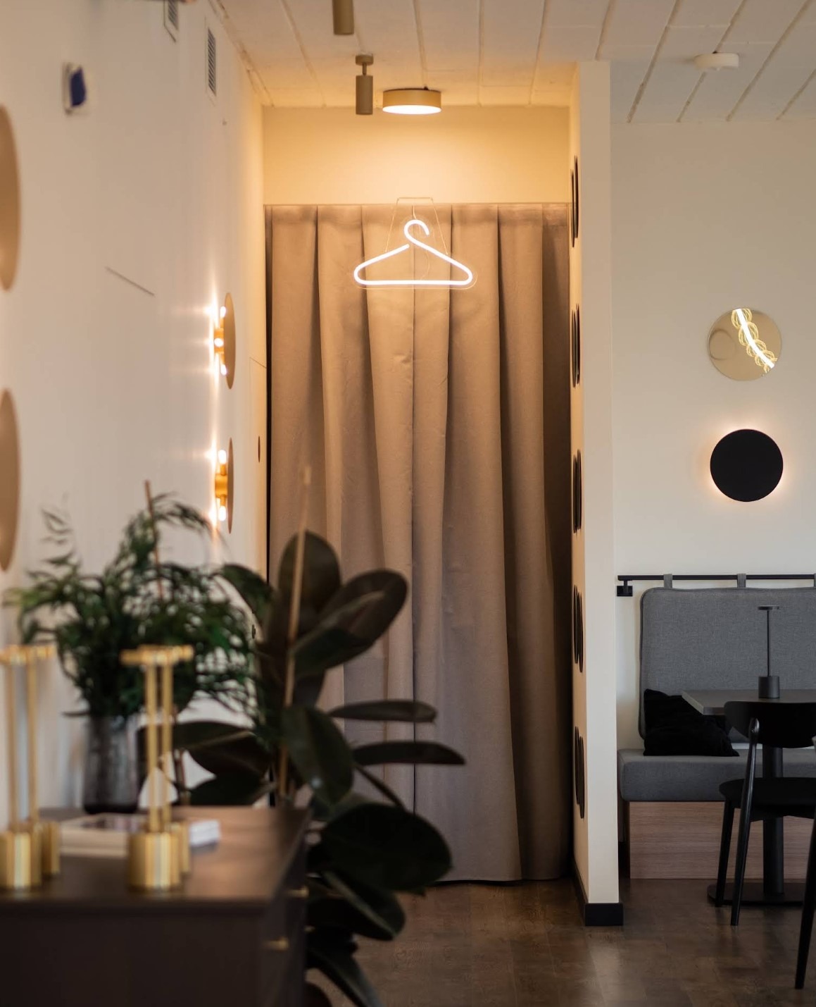 Klimatyczne wnętrze z zasłoniętą przebieralnią, nad którą wisi neon w kształcie wieszaka. Dekoracyjne oświetlenie i rośliny dodają charakteru.