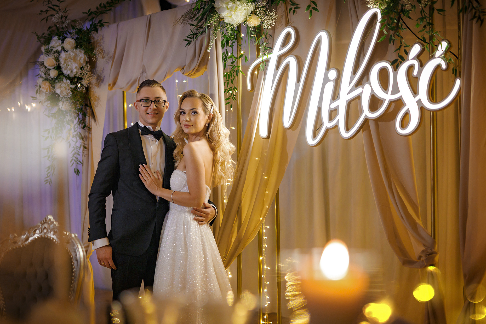 Para młoda w eleganckich strojach pozuje na tle dekoracji ślubnej z napisem 'Miłość' podświetlonym ledami, widoczne kwiaty i delikatne oświetlenie.