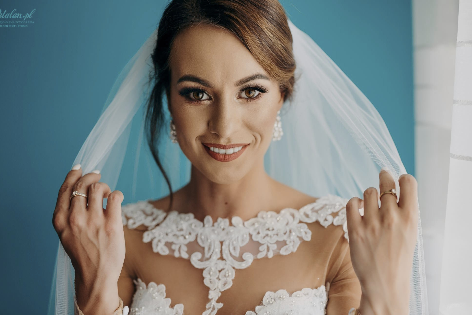 Uśmiechnięta panna młoda w welonie i sukni z koronkowym dekoltem na tle błękitnej ściany, z pierścionkiem zaręczynowym na palcu.