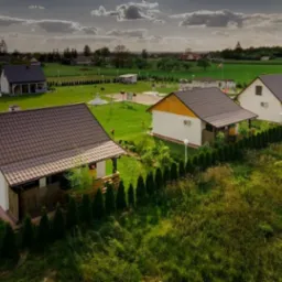 Widok z g&oacute;ry na kompleks domk&oacute;w letniskowych z brązowymi dachami, otoczonych zielenią, z placem zabaw w tle i zachodzącym słońcem.