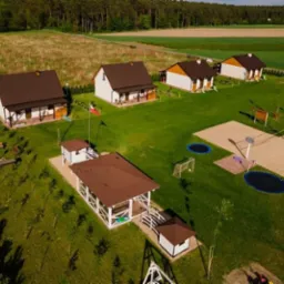 Widok z lotu ptaka na kompleks domków letniskowych z placem zabaw, trampolinami i boiskiem do siatkówki plażowej, otoczony zielenią i polami.