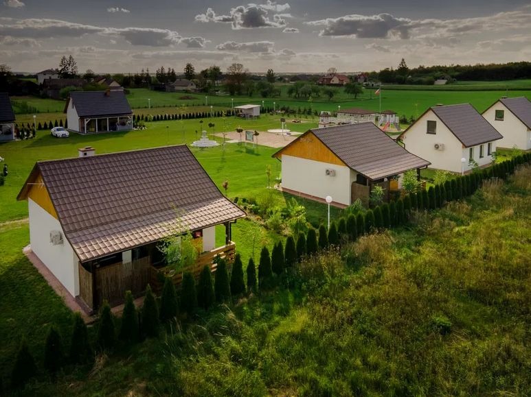 Widok z góry na kompleks domków letniskowych z brązowymi dachami, otoczonych zielenią, z placem zabaw w tle i zachodzącym słońcem.