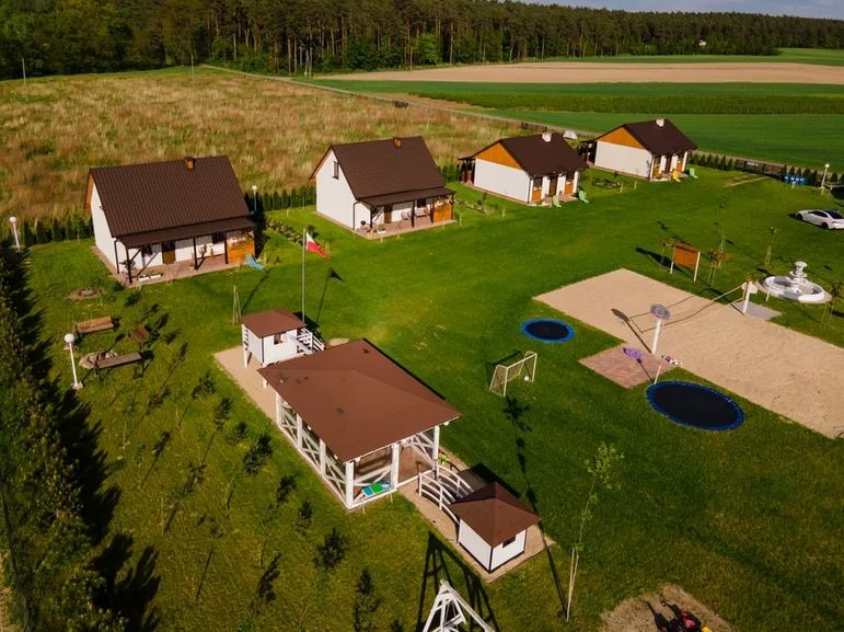 Widok z lotu ptaka na kompleks domków letniskowych z placem zabaw, trampolinami i boiskiem do siatkówki plażowej, otoczony zielenią i polami.