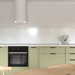 Margareta Gulczyńska GUMA Studio Projektowania Wnętrz - Minimalistyczna kuchnia w odcieniach zieleni z nowoczesnym okapem i sprzętem AGD. Blat z subtelnym wzorem, drewniany stół w tle. Ujęcie podkreśla prostotę i funkcjonalność.
