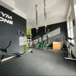 Jasne wnętrze siłowni z czarną ścianą z napisem 'GYM ZONE', sprzętem do ćwiczeń jak wioślarz, sztanga, hantle, drabinki, piłki i ringi gimnastyczne.