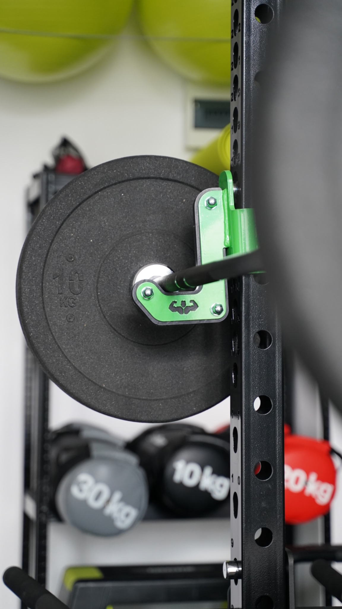 Ciężar 10 kg na gryfie przymocowanym do zielonego uchwytu na czarnej klatce do ćwiczeń, w tle piłki lekarskie na półce.