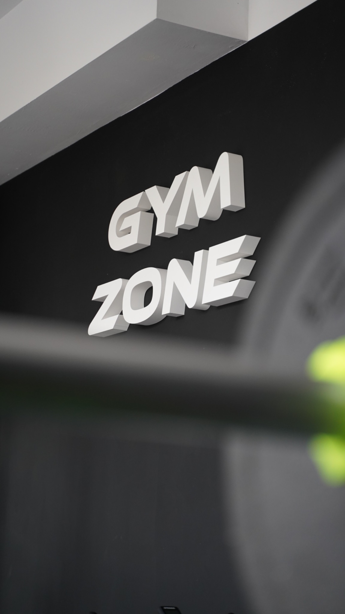 Biały, trójwymiarowy napis 'GYM ZONE' na czarnej ścianie w rogu pomieszczenia z fragmentem białego sufitu i zieloną rośliną w tle.