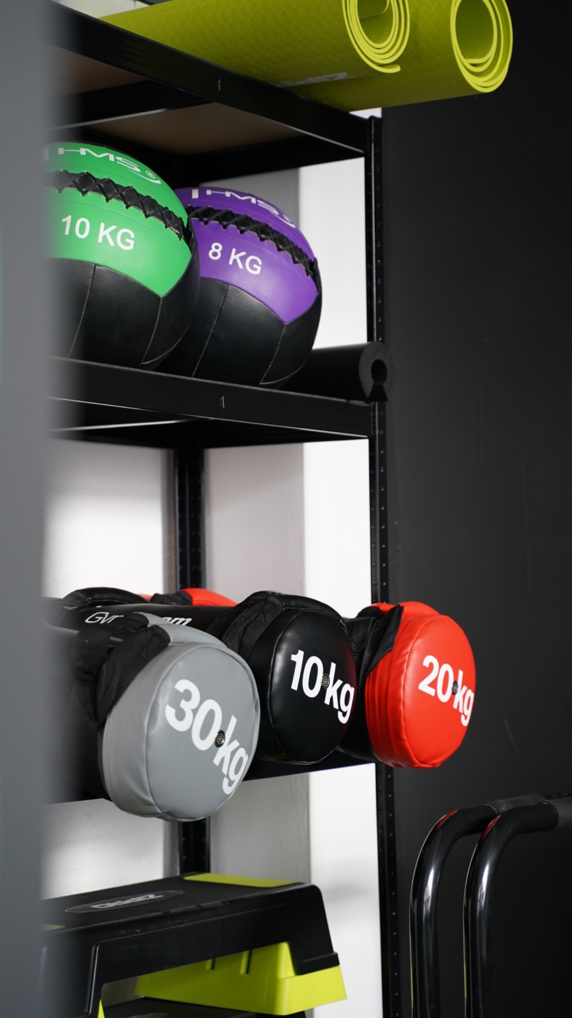 Regał z piłkami lekarskimi o różnych wagach (8, 10, 20, 30 kg) i kolorach (zielony, fioletowy, czarny, szary, czerwony) oraz zwinięte maty do ćwiczeń na górnej półce, widoczny step do ćwiczeń.