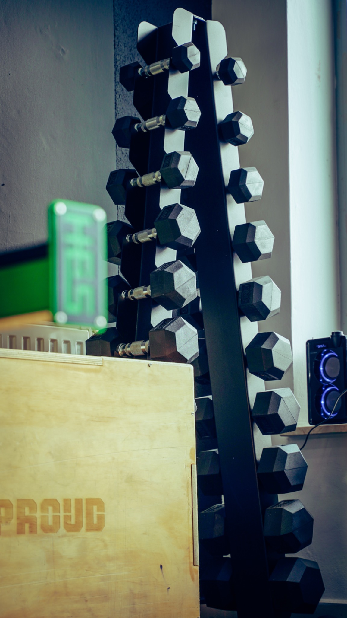 Stojak z czarnymi hantlami o dwunastościennej formie, obok drewniana skrzynia z napisem 'PROUD' w siłowni, widoczny głośnik z niebieskim podświetleniem.
