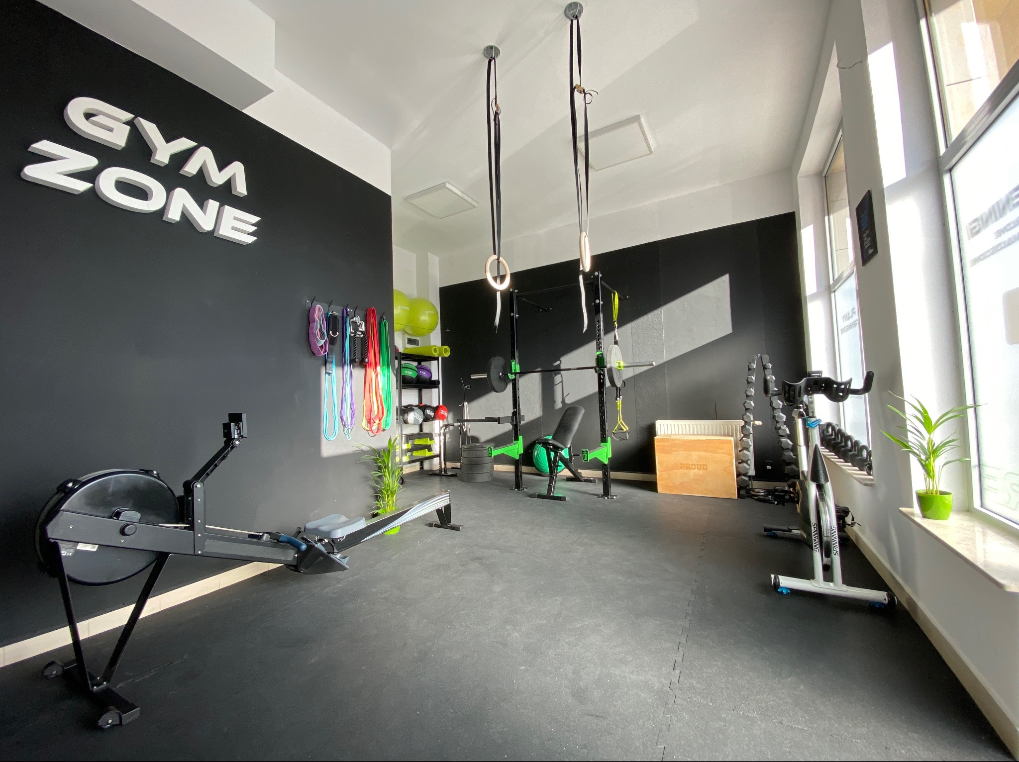 Jasne wnętrze siłowni z czarną ścianą z napisem 'GYM ZONE', sprzętem do ćwiczeń jak wioślarz, sztanga, hantle, drabinki, piłki i ringi gimnastyczne.