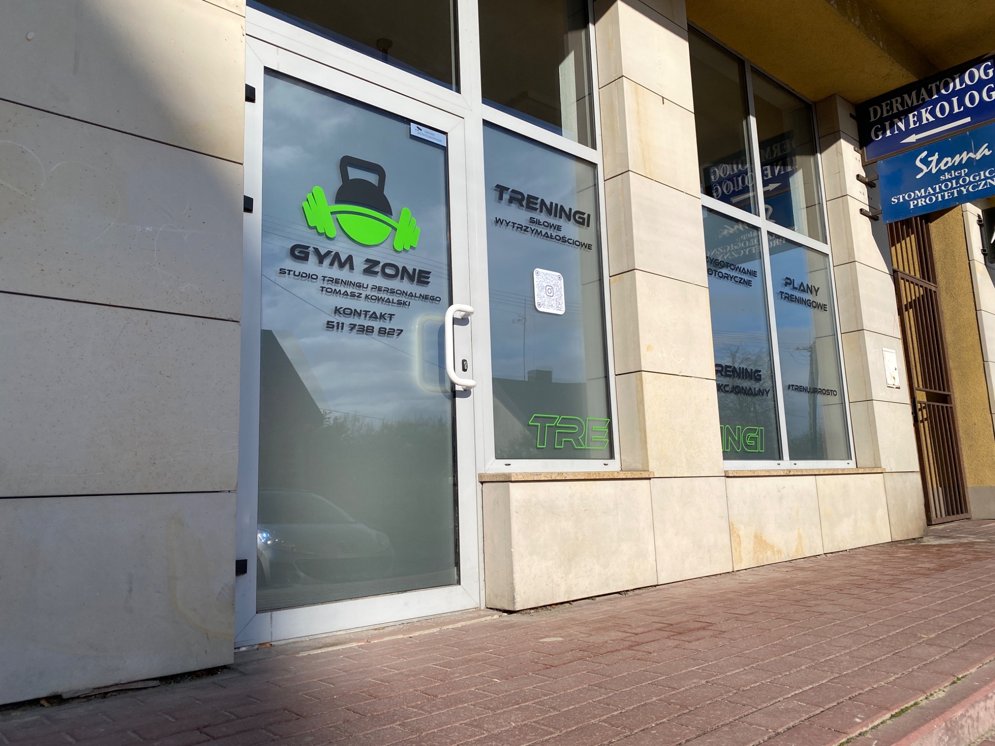 Wejście do studia treningu personalnego Gym Zone z logo z zielonymi hantlami i czarnym kettlebellem na szybie, obok szyld dermatologa i ginekologa.