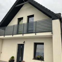 Dwukondygnacyjny dom z kremową elewacją i ciemnoszarą, poziomą balustradą balkonową, widoczny z dołu pod kątem, z czarną stolarką okienną i drzwiami wejściowymi, oraz z roślinami w donicach przy...