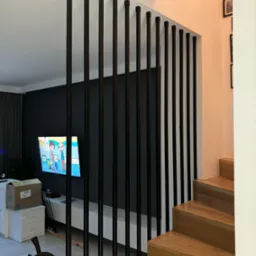 Nowoczesna balustrada schodowa z pionowych, czarnych prętów o kwadratowych zakończeniach, oddzielająca schody od salonu z telewizorem wyświetlającym bajkę.