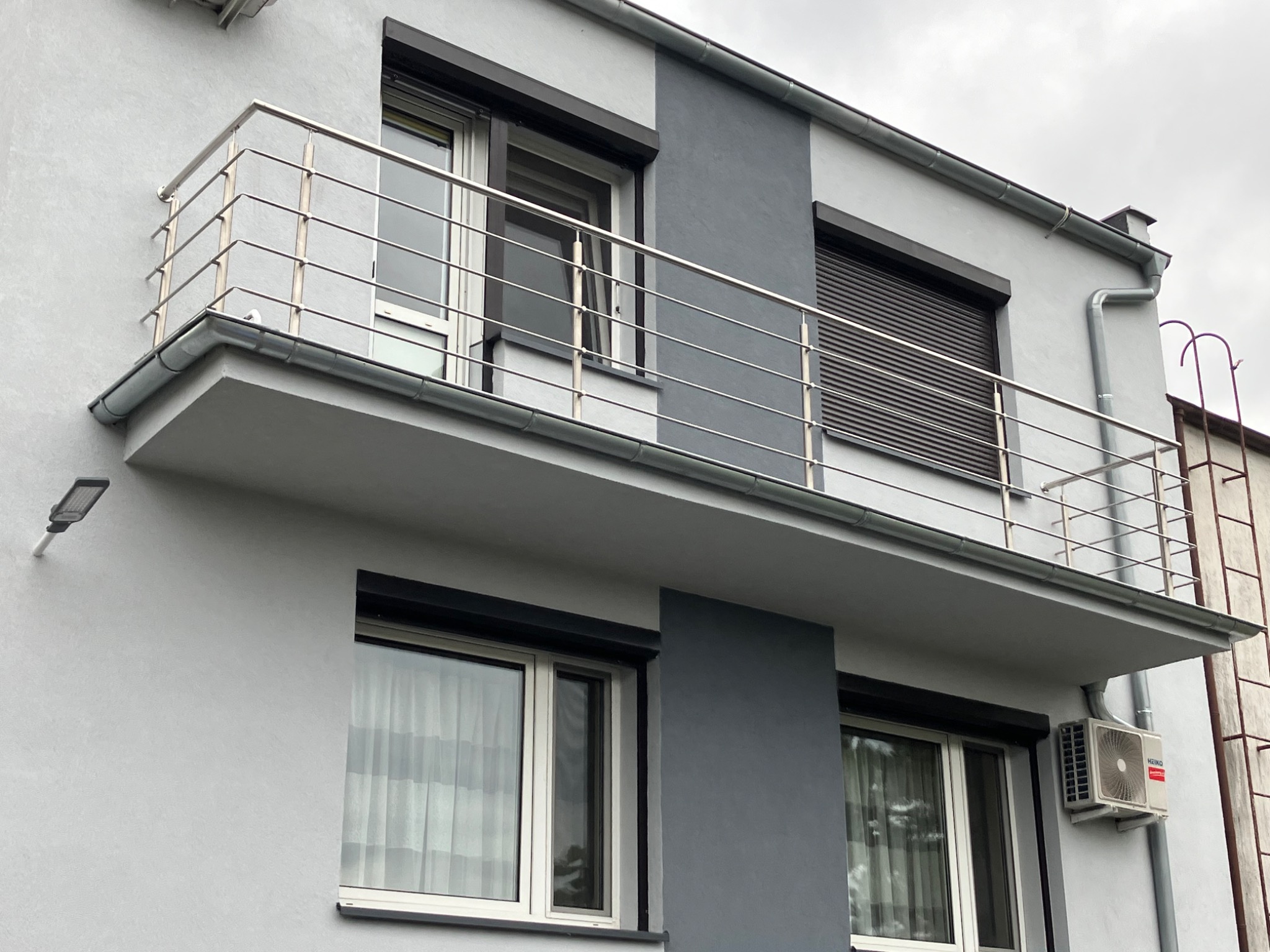 Nowoczesna balustrada nierdzewna z poziomymi prętami na balkonie budynku w odcieniach szarości, widoczna klimatyzacja i rolety zewnętrzne.