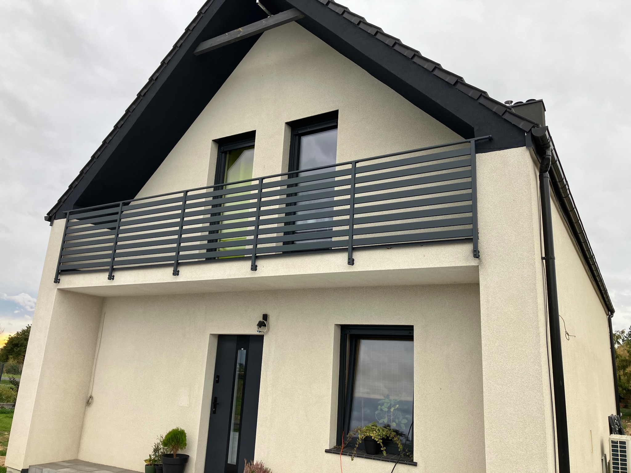 Dwukondygnacyjny dom z kremową elewacją i ciemnoszarą, poziomą balustradą balkonową, widoczny z dołu pod kątem, z czarną stolarką okienną i drzwiami wejściowymi, oraz z roślinami w donicach przy...