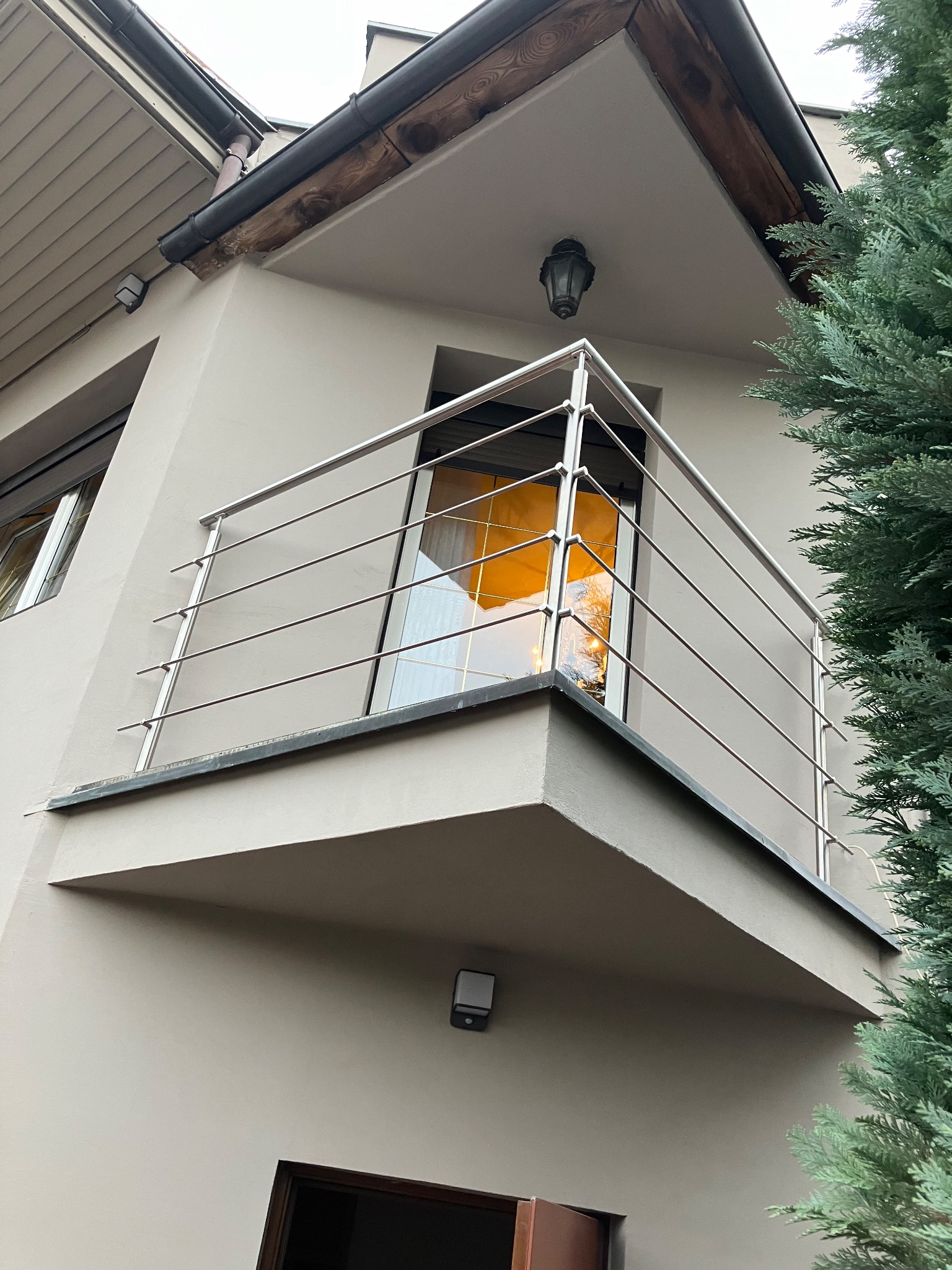 Balkon z nowoczesną, minimalistyczną balustradą ze stali nierdzewnej, widok z dołu, z fragmentem iglastego drzewa po prawej stronie. W oknie balkonu odbija się ciepłe, pomarańczowe światło.