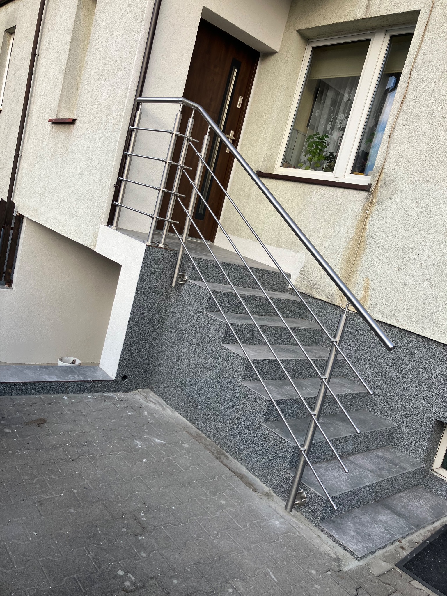 Zewnętrzne schody z nowoczesną balustradą ze stali nierdzewnej w Stęszewie, prowadzące do drzwi wejściowych. Widoczne szare płytki schodowe i elewacja budynku o jasnej strukturze.