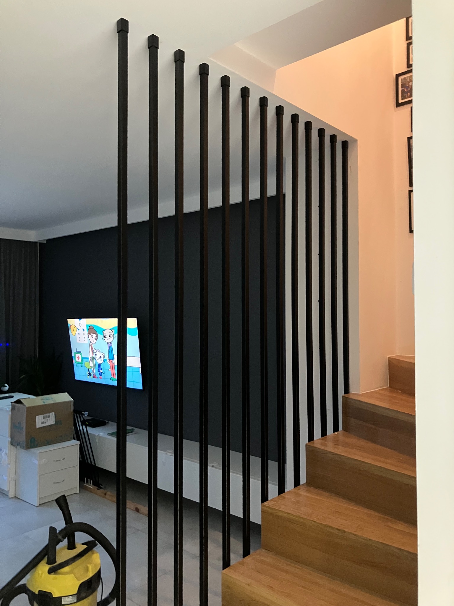 Nowoczesna balustrada schodowa z pionowych, czarnych prętów o kwadratowych zakończeniach, oddzielająca schody od salonu z telewizorem wyświetlającym bajkę.