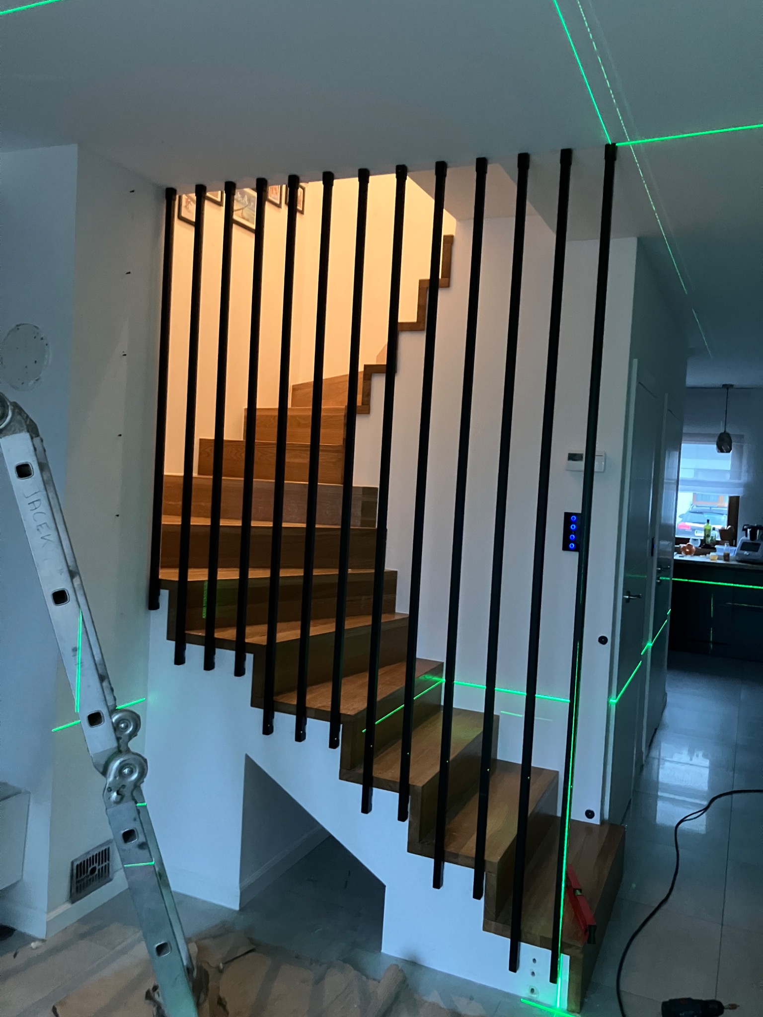 Drewniane schody z czarnymi, pionowymi balustradami zabezpieczającymi, oświetlone zielonym laserem poziomującym, widoczne narzędzia i drabina.