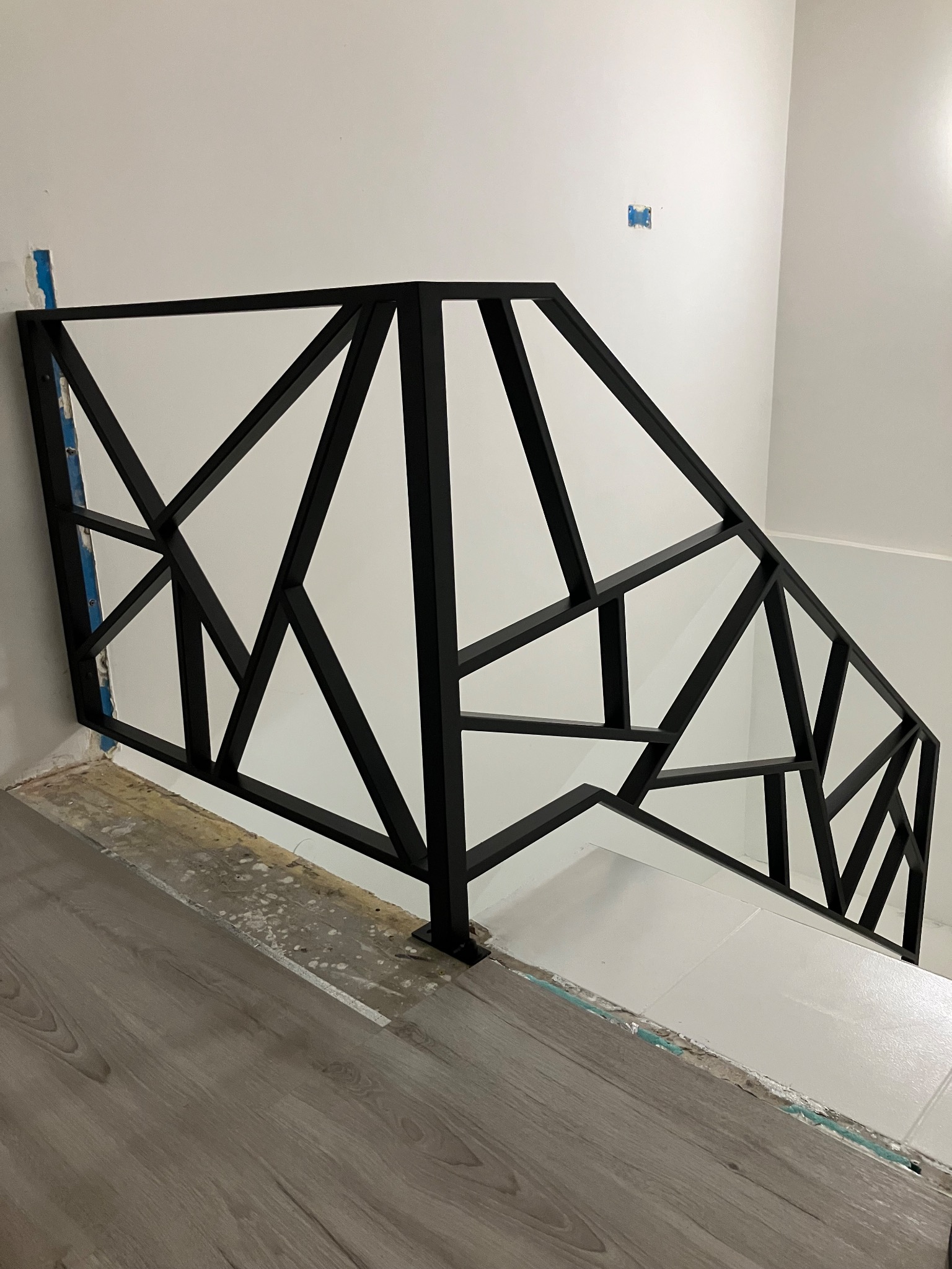 Czarna balustrada schodowa o geometrycznym wzorze, wykonana z metalowych profili, zamontowana na białej ścianie i jasnych schodach, widoczne niedokończone wykończenie podłogi.