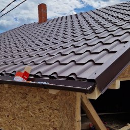 Igor Lysenko - Montaż brązowego dachu z blachodachówki modułowej, widoczne elementy konstrukcyjne, komin z cegły w tle i fragment błękitnego nieba z chmurami.