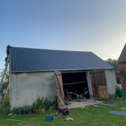 Igor Lysenko - Montaż blachy na dachu starego budynku gospodarczego; na drabinie dekarz, otwarte wrota, narzędzia na trawie.