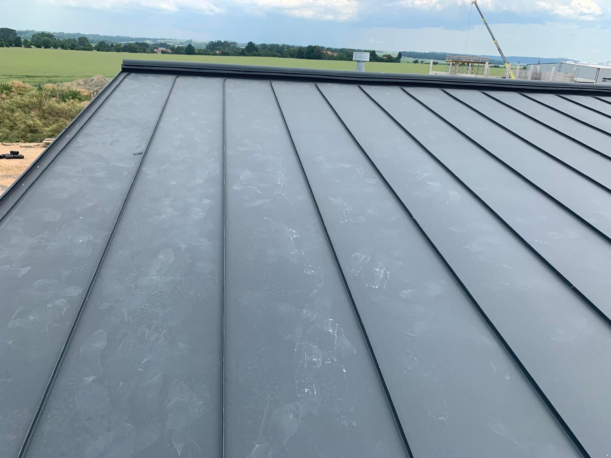 Nowy, szary dach z blachy na rąbek stojący, widoczne ślady butów dekarza, w tle zielone pole i budowa z dźwigiem.