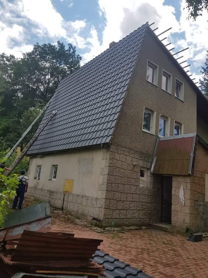 Dom z nowym, ciemnoszarym dachem z dachówki, widoczna drabina oparta o dach i fragment pracownika na drabinie, na pierwszym planie fragmenty starych pokryć dachowych