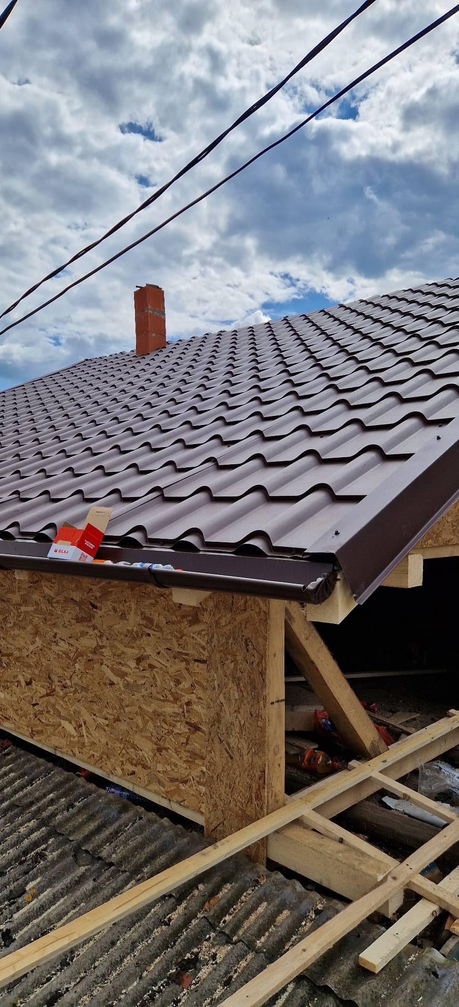 Montaż brązowego dachu z blachodachówki modułowej, widoczne elementy konstrukcyjne, komin z cegły w tle i fragment błękitnego nieba z chmurami.