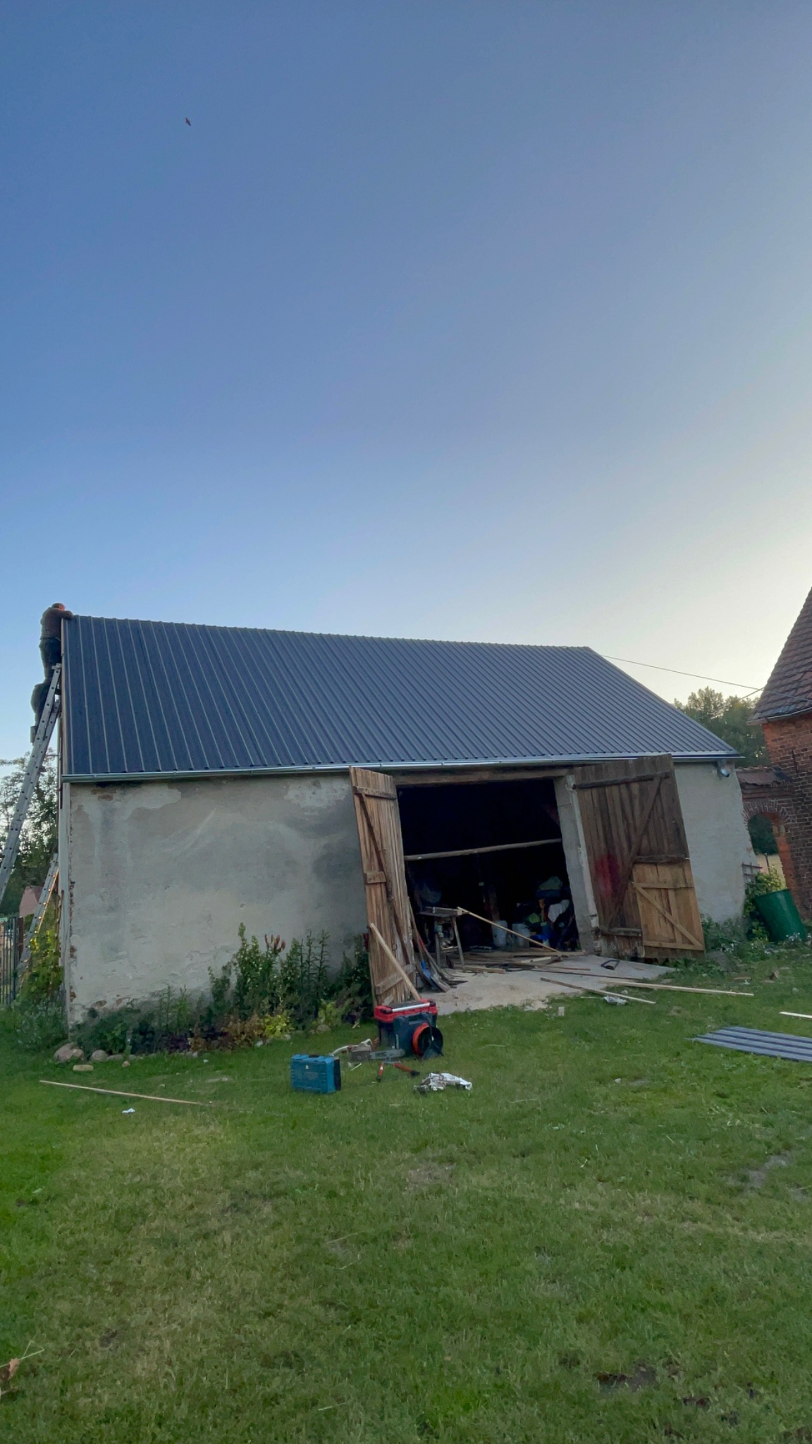 Montaż blachy na dachu starego budynku gospodarczego; na drabinie dekarz, otwarte wrota, narzędzia na trawie.