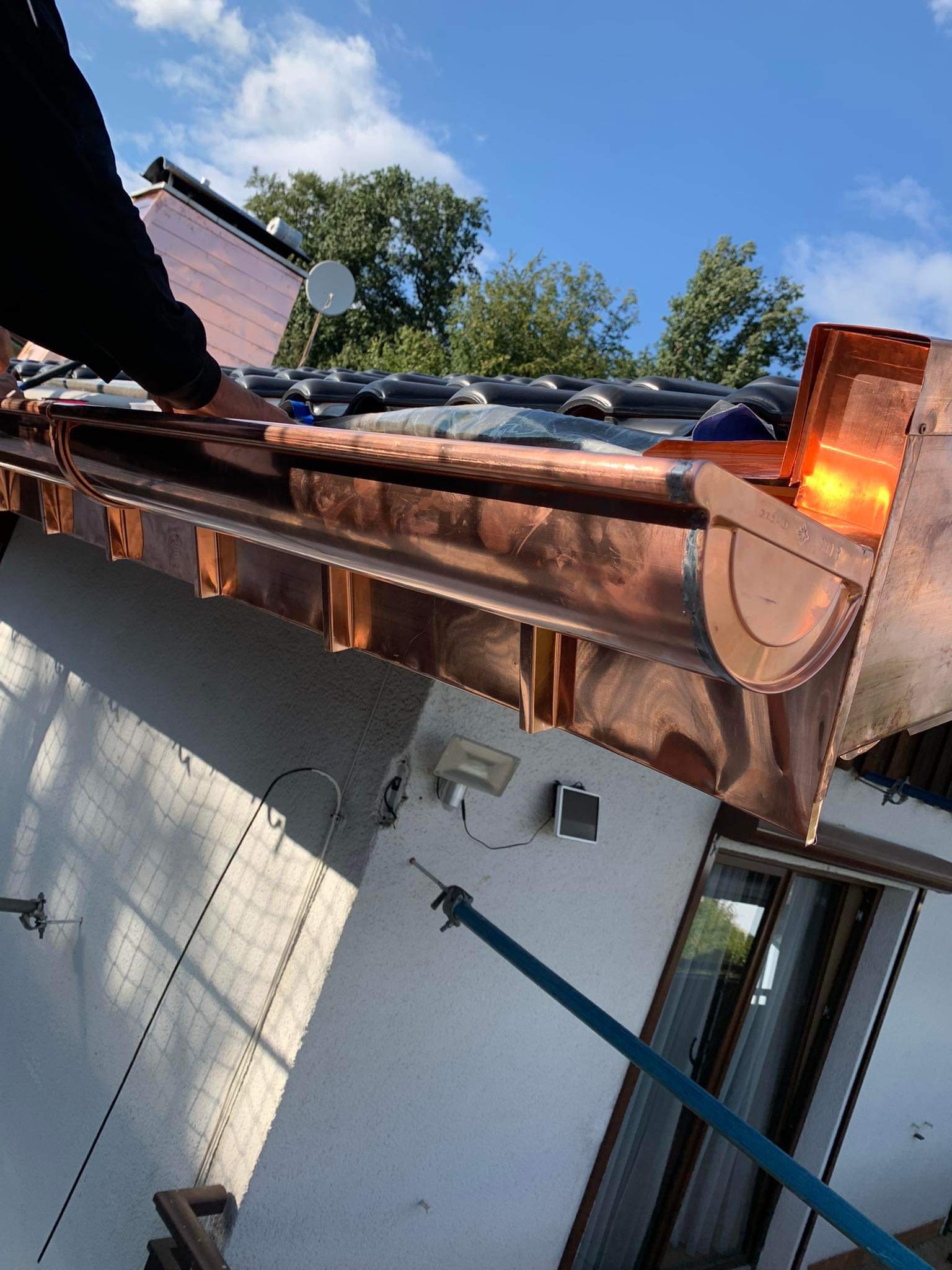 Miedziana rynna dachowa, montowana przez dekarza na tle błękitnego nieba i zielonych drzew, widoczne narzędzia i elementy mocujące.