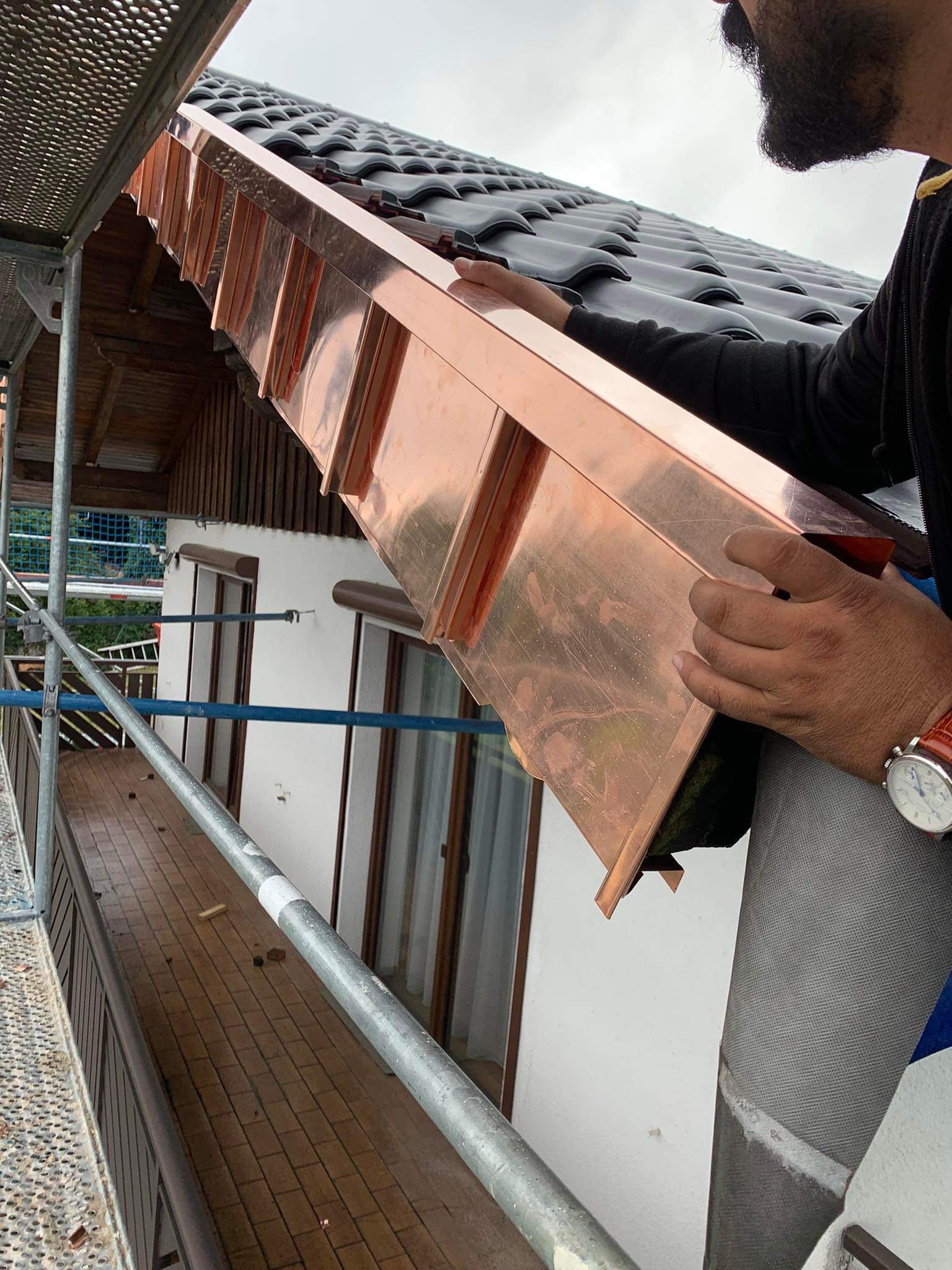 Dekarz montuje miedzianą obróbkę blacharską na krawędzi dachu z czarnej dachówki, widoczne rusztowanie i fragment elewacji budynku.