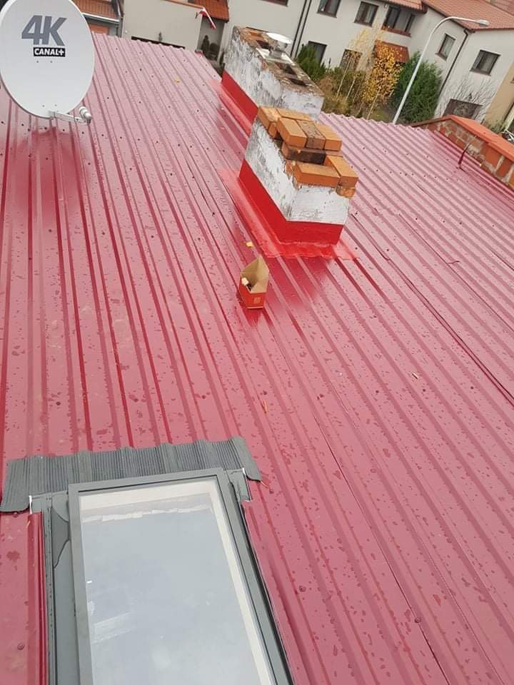 Czerwony dach z blachy trapezowej, widoczny komin z uszkodzonymi cegłami i świeżym czerwonym uszczelnieniem, okno dachowe oraz antena satelitarna 4K, lekko wilgotna powierzchnia.
