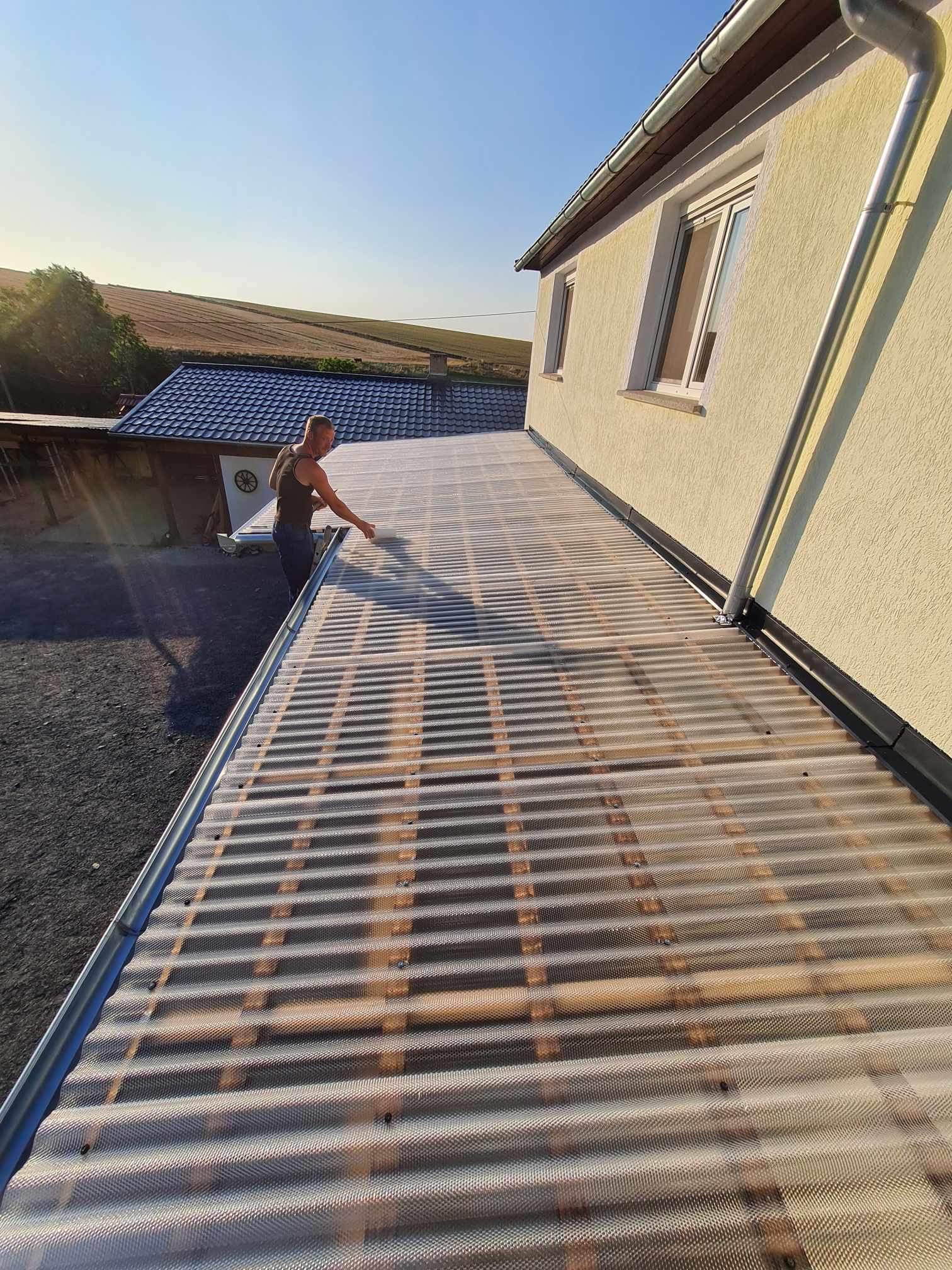 Pracownik montuje przezroczyste pokrycie dachowe z tworzywa sztucznego na konstrukcji drewnianej we Wrocławiu, widoczny fragment elewacji budynku i oddalone pola uprawne.